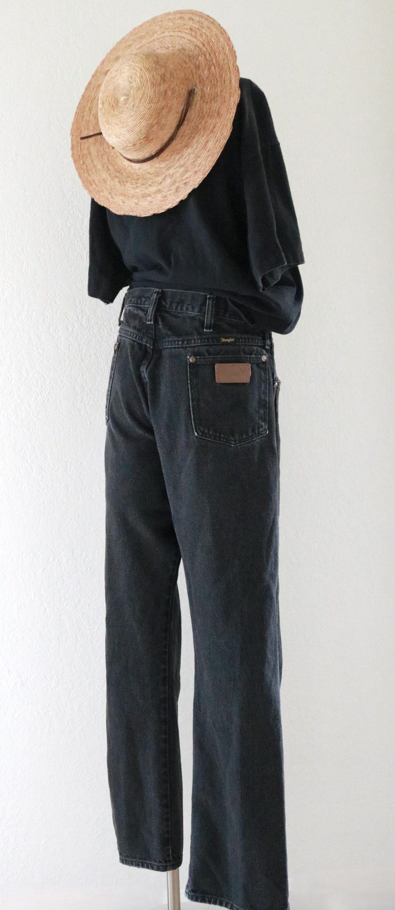 May include: A person wearing a black t-shirt and dark wash denim jeans. The jeans have a brown leather patch on the back pocket. A straw hat with a brown band is worn.