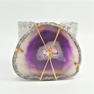 May include: A polished agate slice with a vibrant purple center and a white outer edge. The stone is wrapped with gold-colored wire in a crisscross pattern. The agate is a natural stone, and the wire adds a decorative element.