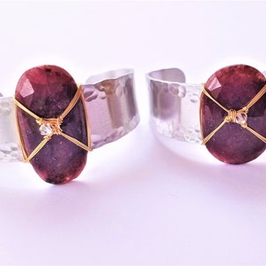 May include: Two silver-tone cuff bracelets with a hammered texture. Each bracelet features a large, oval, faceted, deep red gemstone secured with gold-colored wire. A small, clear crystal is also incorporated into the design.