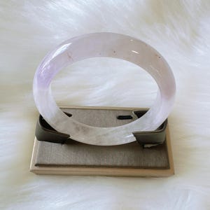 May include: A translucent, lavender and white gemstone bangle bracelet. The bracelet is displayed on a gray and brown stand against a white, fluffy background. The bracelet has a smooth, polished surface.