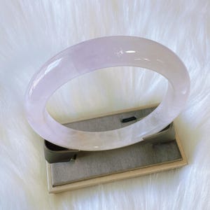 May include: A translucent, lavender-colored jadeite bangle bracelet. The smooth, polished surface reflects light, showcasing the natural variations in the stone. The bracelet is displayed on a gray and beige stand against a white, fluffy background.