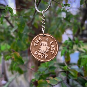 Puede incluir: Un llavero de madera con un diseño de jarra de cerveza y el texto "The Last Drop".