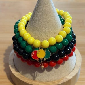 May include: A stack of beaded bracelets in red, black, green, and yellow. A gold charm with red, yellow, and green balloons hangs from the top bracelet. The bracelets are displayed on a beige cone-shaped stand.