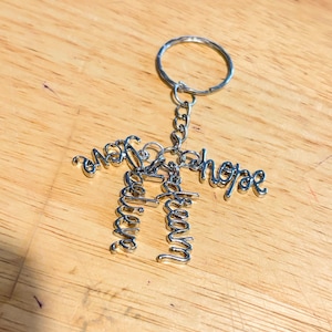 May include: A silver-colored metal keychain with the words "love", "dream", and "hope" in a cursive font. The keychain has a ring and chain attachment. The words are connected to form a figure.