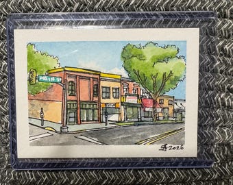 Puede incluir: Una acuarela de una esquina con edificios de ladrillo, una señal de calle verde que dice MAIN ST y un gran árbol verde. La obra presenta contornos de tinta negra, un cielo azul y está firmada con el año 2026 en la esquina inferior derecha.