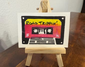 Puede incluir: Una pequeña pintura rectangular en acuarela de un casete sobre un caballete de madera. El casete presenta un fondo degradado en amarillo y rosa con el texto ROAD TRIPPIN' en tinta negra, carretes blancos y detalles de rayas blancas y negras.