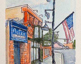 Puede incluir: Una acuarela de una escena callejera en Moab, UT, que muestra un edificio de ladrillo con un letrero azul que dice Alley Clothing, una farola negra alta y una bandera estadounidense colgando. La obra está firmada y fechada en 2026.