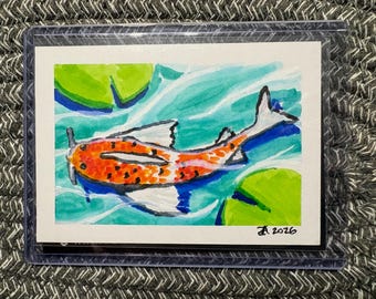 Puede incluir: Una pintura de acuarela de un pez koi naranja y blanco con manchas negras nadando en agua azul con nenúfares verdes. La obra está dentro de una funda protectora de plástico transparente y presenta la firma manuscrita JA 2026.