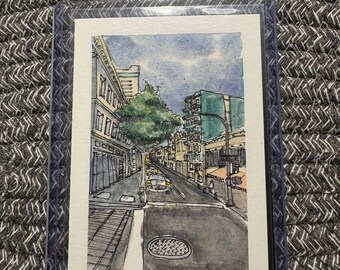 Puede incluir: Una pintura en acuarela de una escena de calle urbana con edificios, un gran árbol verde y coches en la carretera. La obra incluye una tapa de alcantarilla en primer plano y está firmada con el texto 2026 en la esquina inferior derecha.