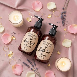 Puede incluir: Dos botellas de loción marrones con bombas negras, etiquetadas como "Love Spell" y "Lush Ice", están sobre una superficie rosa con velas, pétalos de rosa y detalles dorados. Cada botella contiene 237 ml de loción.