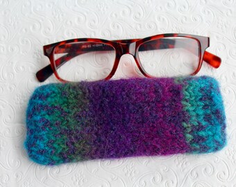 May include: A pair of tortoiseshell-patterned eyeglasses rests atop a hand-knitted glasses case. The case is a blend of blue, green, and purple hues. The glasses have a rectangular frame.