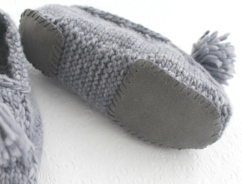 Puede incluir: Un par de zapatillas de punto grises con un pomp&oacute;n en una zapatilla. Las zapatillas tienen una suela de goma negra.