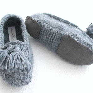 Puede incluir: Un par de zapatillas de punto grises con un detalle de borla y una suela de goma negra.