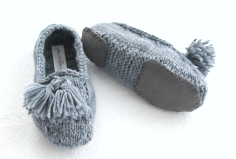Puede incluir: Un par de zapatillas de punto grises con un detalle de borla y una suela de goma negra.