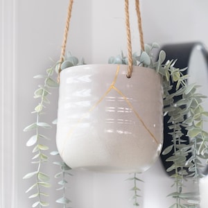 Könnte beinhalten: Ein weißer Keramik-Hängepflanzentopf mit goldenen Akzenten, aufgehängt an einem Naturseil. Der Topf ist mit künstlichem, grün-grauem Eukalyptus gefüllt. Ein dekoratives Element für die Inneneinrichtung.