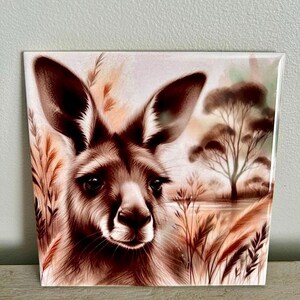 May include: A close-up of a brown kangaroo's face with large ears and a soft, furry muzzle. The kangaroo is looking directly at the viewer with a gentle expression. The background is a soft, blurred image of tall grass and a distant tree.