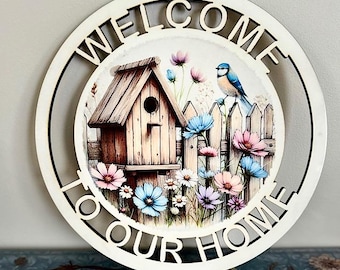 May include: A white wooden round sign with the words "Welcome to Our Home" in a black cutout font. The sign features a colorful image of a birdhouse, a bluebird, and flowers in front of a white picket fence.