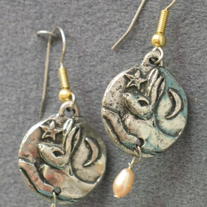 May include: A pair of silver earrings with a rabbit and crescent moon design. Each earring has a small pearl hanging from it.