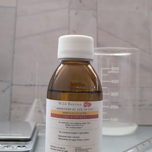 May include: A brown glass bottle with a white cap and a label reading Wild Berries, Ambroxan al 10% en DPG, Ambroxide (dilución), CAS 6790-58-5, and Contenido neto: 100 g. The bottle is positioned near a graduated beaker and a digital scale.