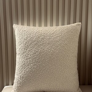 May include: A square, cream-coloured cushion with a textured, bobbled surface. The cushion has a neutral tone and is likely for decorative use. The background is a light beige wall with vertical, ridged lines.