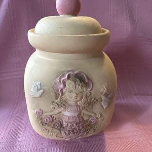Może przedstawiać: Ceramiczny słoik w kremowym kolorze z pokrywką. Słoik posiada relief przedstawiający dziewczynkę w różowej sukience i kapeluszu, otoczoną różami i motylami. Pokrywka zwieńczona jest różową kulą.