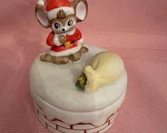 May include: A ceramic Christmas figurine featuring a mouse dressed in a Santa suit and hat. The mouse sits atop a white chimney-shaped box with a brick design. A yellow sack with a green item peeks out.