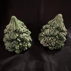 Może przedstawiać: Dwie zielone solniczki i pieprzniczki ceramiczne w kształcie choinek. Każde drzewko ma teksturowany, liściasty wzór i małe otwory na górze do dozowania. Solniczki i pieprzniczki są ustawione na ciemnym tle.