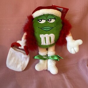 Puede incluir: Un personaje de caramelo verde de peluche que lleva un gorro de Papá Noel con ribete blanco y ribete de plumas rojas. El personaje tiene una "M" blanca en el frente, guantes blancos y botas blancas con cinta verde y una pequeña bolsa blanca.