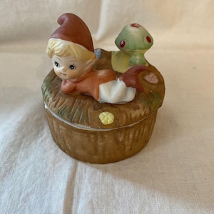 Puede incluir: Una caja de baratijas de cerámica con un diseño caprichoso. La caja marrón tiene una tapa, y una pequeña figura de gnomo está acostada encima. Una seta verde con lunares rojos también está en la tapa. El gnomo lleva un sombrero rojo y un atuendo naranja.