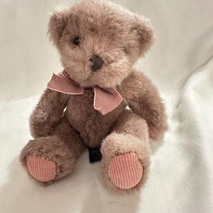 Puede incluir: Un pequeño oso de peluche en tonos marrones y rosas. El oso tiene un lazo rosa alrededor del cuello y almohadillas rosas, tipo pana, en los pies. El oso está sentado sobre una superficie blanca.