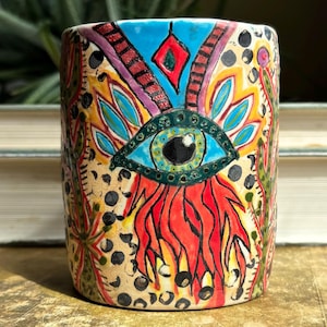 May include: A hand-painted ceramic cup with an eye design. The cup features a blue eye with green details, red flames, and a blue and red diamond shape. The background is beige with black dots and colorful floral patterns.