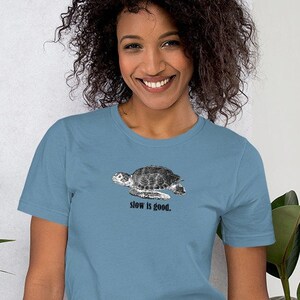Puede incluir: Una camiseta azul claro con una ilustración en blanco y negro de una tortuga marina y el texto "slow is good."