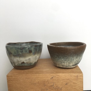 May include: Two ceramic bowls with a brown, green, and white glaze. The bowls are sitting on a wooden surface.