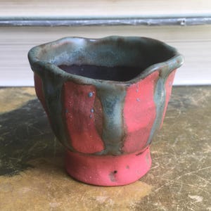 May include: A handcrafted ceramic cup with a distinctive design. The cup has a red base and sides with vertical, irregular stripes. The rim and upper section are a contrasting green-blue colour. Small blue speckles add detail to the surface.