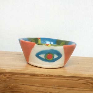 May include: A small, hand-painted ceramic bowl with a blue eye design. The bowl is decorated with green, orange, and yellow accents.