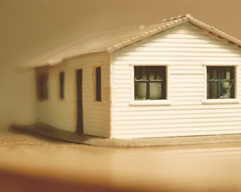 May include: A white miniature house with a brown roof, two windows with black frames, and a door. The house is sitting on a brown surface.