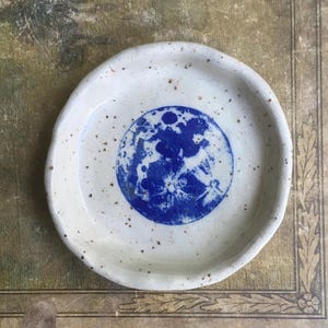 May include: A white ceramic dish with a blue and white moon design. The dish has a speckled glaze and a slightly irregular shape.