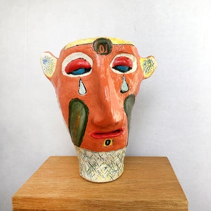 May include: A ceramic sculpture of a face with a yellow, orange, red, blue, green, and white paint design. The face has a large nose and is crying. The sculpture is on a wooden surface.