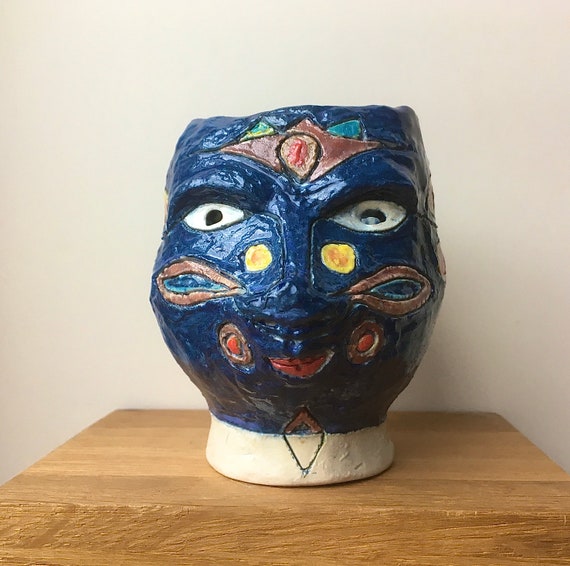 A small primitive ceramic head sculpture with raw, ancient-artifact energy — handmade pottery that looks like it belongs in an archaeology dig.