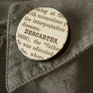 May include: A round button with the text "DESCARTES, 1650" printed on a vintage paper background. The button is set against a gray fabric background, likely a shirt or jacket.