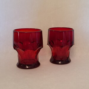 May include: Two red glass tumblers with a faceted design. The glasses are empty and sitting on a white surface.