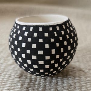May include: A small, round ceramic pot with a black exterior patterned with white squares. The interior is white. The pot sits on a textured, light-colored surface. The design is a modern, geometric pattern.