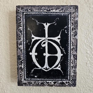 Könnte beinhalten: Eine rechteckige schwarz-weiße dekorative Fliese mit einem verzierten Rahmen. Die Fliese zeigt ein weißes, stilisiertes Monogramm oder Emblem vor einem schwarzen Hintergrund. Der Rahmen hat ein detailliertes Design im Vintage-Stil.