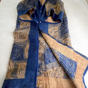May include: A reversible, long, sleeveless wrap garment featuring a fuzzy, high-pile texture. One side displays a dark blue and tan leaf pattern, while the other side is solid tan. The edges are trimmed in dark blue with silver metal snap buttons.