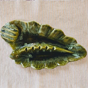May include: A green ceramic ashtray shaped like a leaf with a small, separate compartment for cigarettes. The ashtray has a glossy finish and a textured surface.