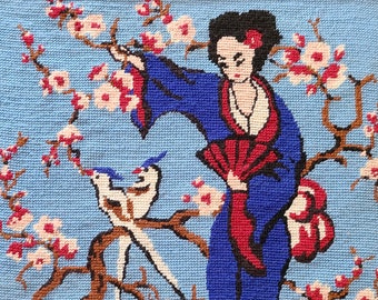 May include: A framed needlepoint embroidery depicting a woman in a blue and red kimono standing in a blooming cherry blossom tree. Two white birds are perched on a branch.
