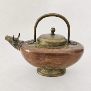 May include: A small, antique copper and brass teapot with a rounded body and a spout shaped like a cow's head. The teapot has a brass handle and lid.