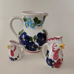 May include: A set of three ceramic pitchers with floral designs. The largest pitcher is white with blue and green flowers. The two smaller pitchers are shaped like roosters and are decorated with blue, pink, and green flowers.