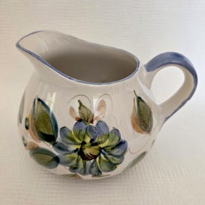 May include: A cream-colored ceramic creamer pitcher featuring a hand-painted floral design in shades of blue, green, and brown. The pitcher has a rounded body, a curved handle, and a blue-trimmed rim, showcasing a classic, functional design.