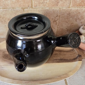 May include: A black ceramic teapot with a lid and a spout. The teapot is sitting on a wooden tray.